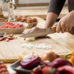 Pessoa cortando alimentos em cima de uma tábua de madeira em bancada de cozinha.
