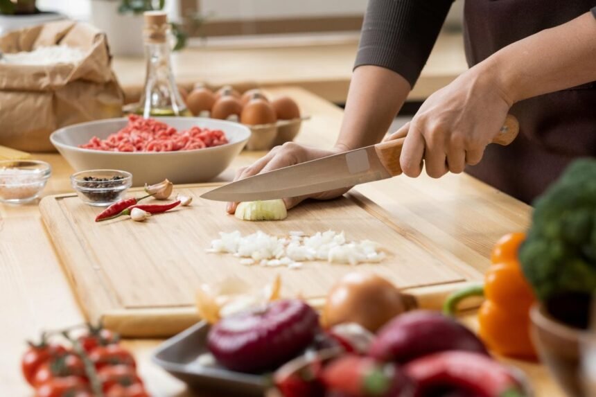Pessoa cortando alimentos em cima de uma tábua de madeira em bancada de cozinha.