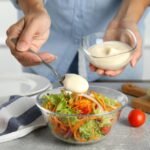 Foto mostra uma pessoa colocando molho na salada em um pote de vidro.