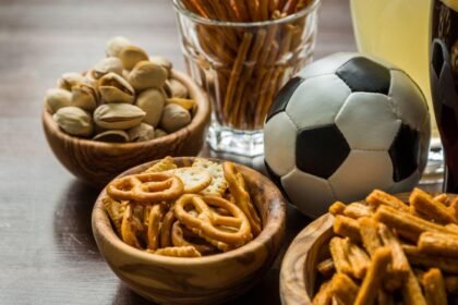 Imagem de petiscos em potes pequenos em cima de uma mesa com uma bola de futebol e um copa grande de refrigerante