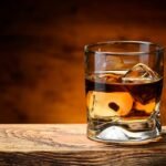 Foto mostra um copo de whisky cheio em cima da mesa de madeira.