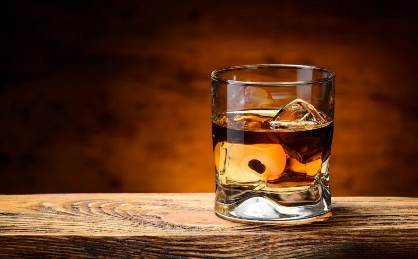 Foto mostra um copo de whisky cheio em cima da mesa de madeira.