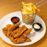 Imagem de iscas de frango fritas e uma porção de batatas frita em um prato com molhos em pequenos potes
