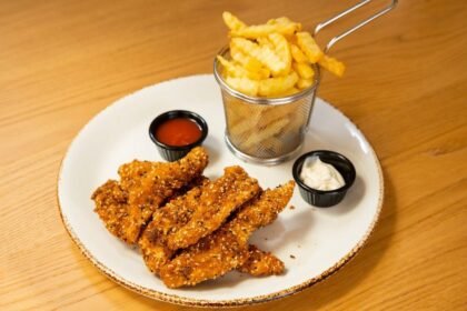 Imagem de iscas de frango fritas e uma porção de batatas frita em um prato com molhos em pequenos potes