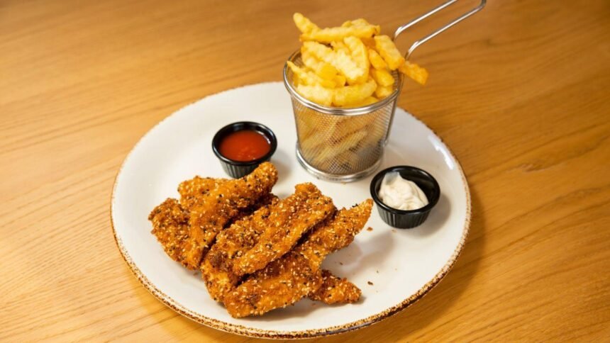 Imagem de iscas de frango fritas e uma porção de batatas frita em um prato com molhos em pequenos potes