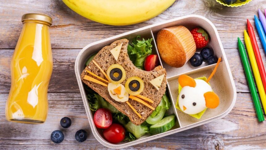 Imagem de uma lancheira com lanche natural em pão de forma com rosto de gatinho, frutas e um suco de laranja em uma garrafa de vidro ao lado