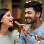 Imagem de um homem dando um pedaço de chocolate na boca de uma mulher com taças de vinho branco na mesa