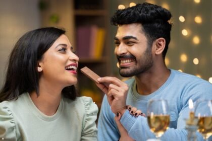 Imagem de um homem dando um pedaço de chocolate na boca de uma mulher com taças de vinho branco na mesa