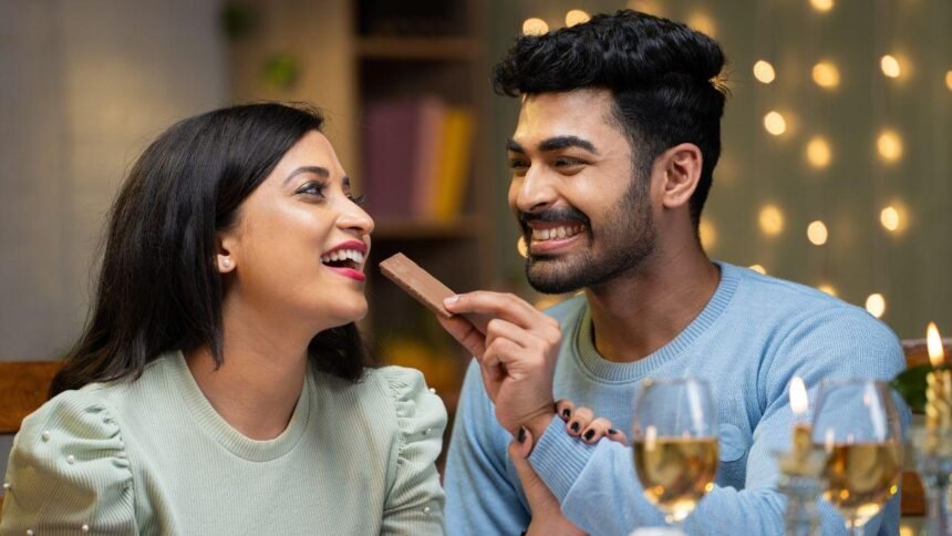 Imagem de um homem dando um pedaço de chocolate na boca de uma mulher com taças de vinho branco na mesa