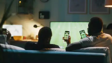 Framestock/ Adobe Stock 13/07/2026 - Bets - African American man using smartphone and clinking bottles of beer with friends while chilling on sofa and watching soccer game on TV. Foto: Framestock/ Adobe Stock